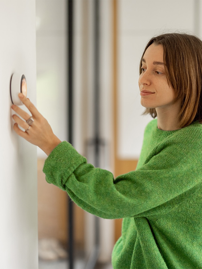 Woman controlling temperature with a smart thermostat mounted on the wall at modern apartment. Concept of smart technologies at domestic life. Woman wearing green sweater