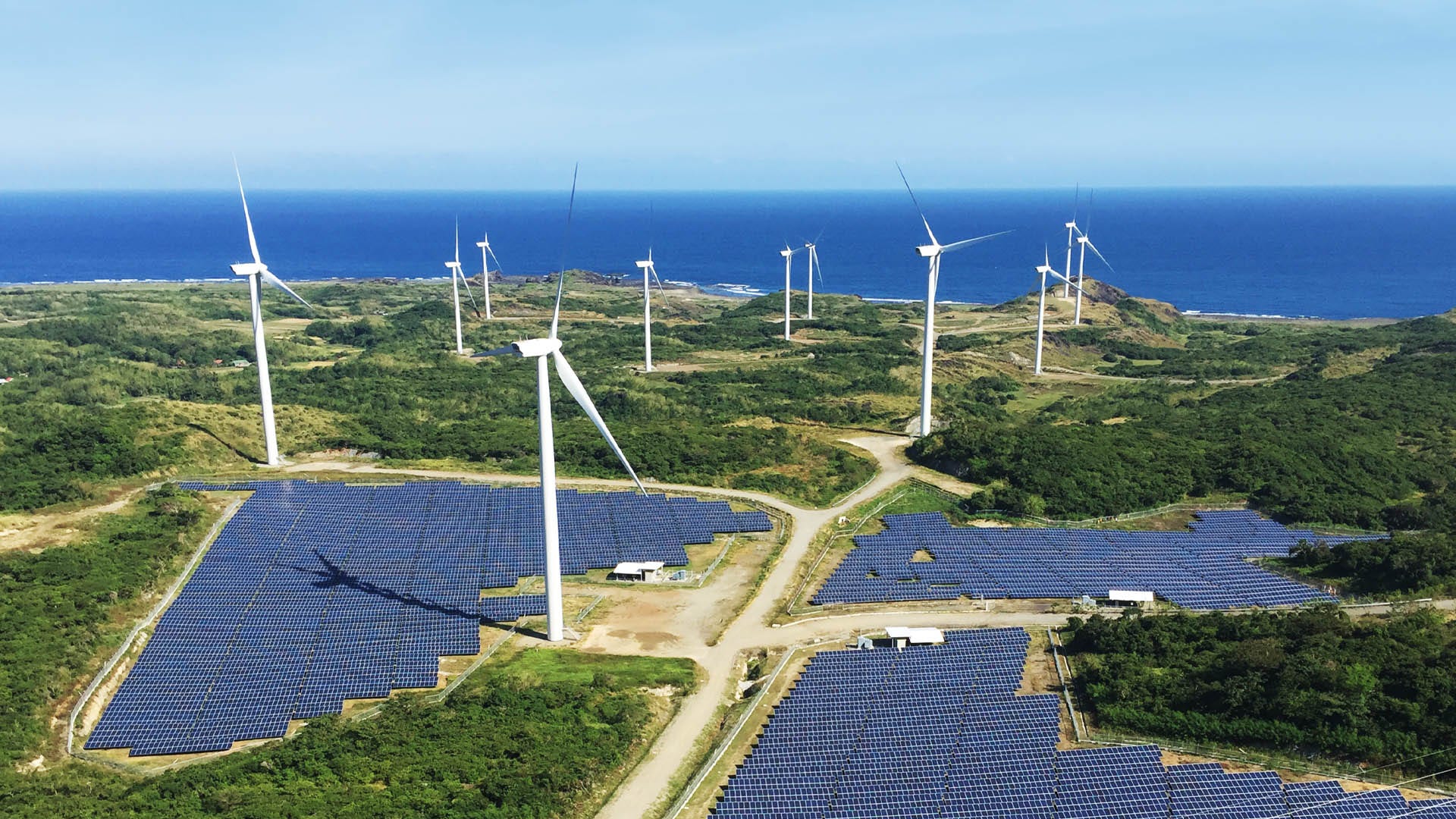 high angle of wind turbines and solar panels with surrounding road, trees and coastline