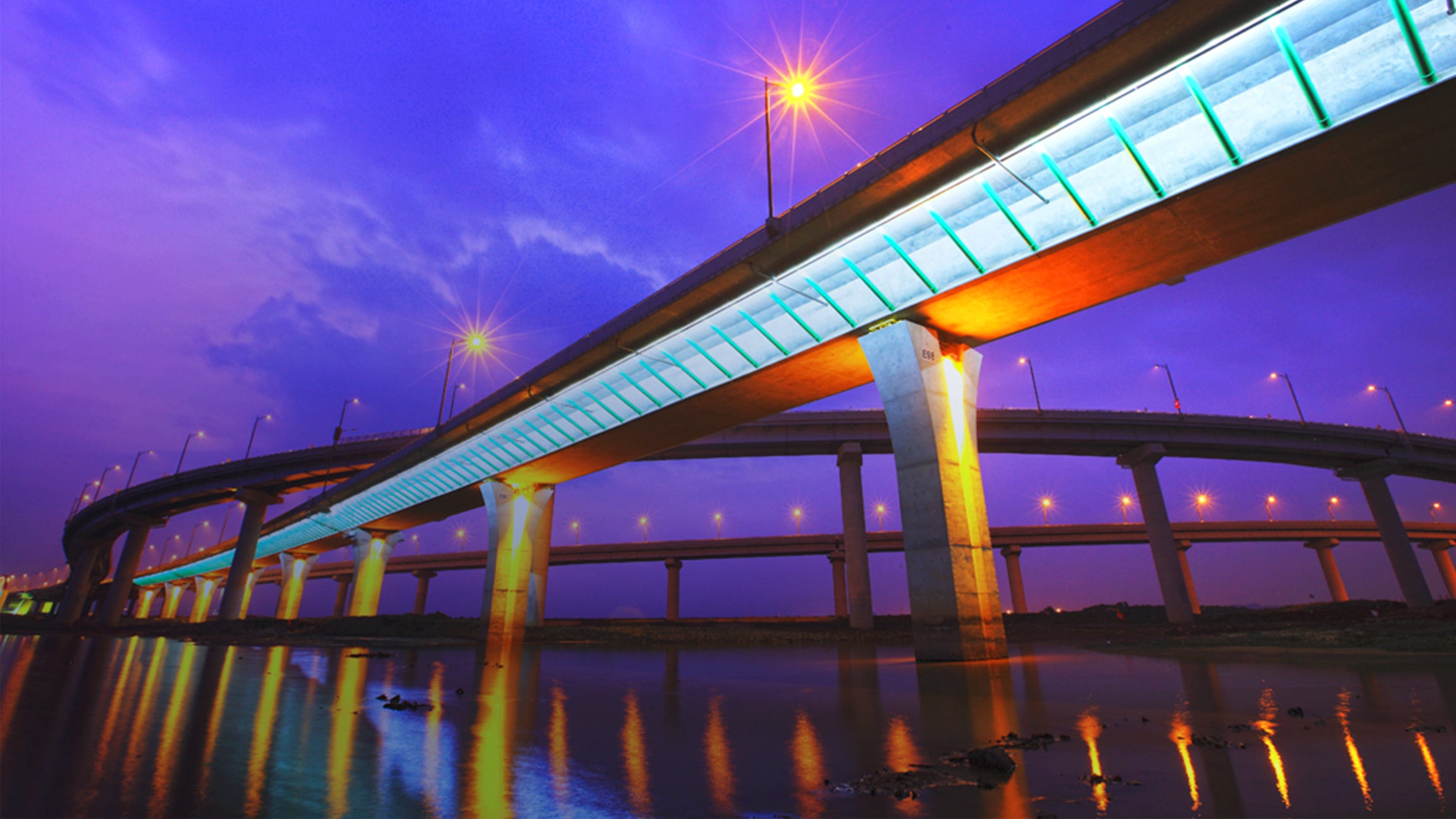 pylon-elevated colourfully-lit roads over water with purple sky in background