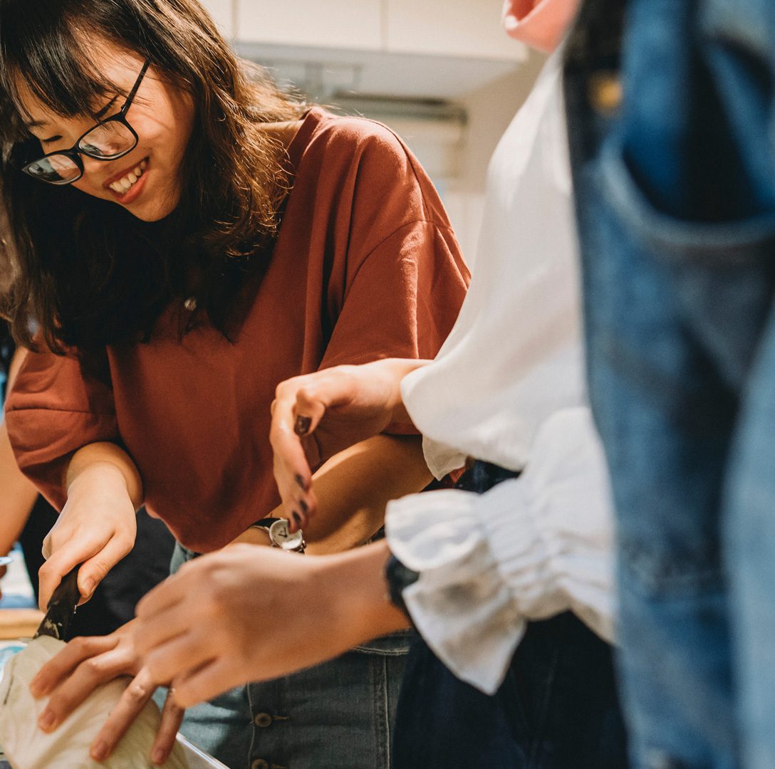 A group of people preparing food together in a kitchen, with one person chopping ingredients at the counter while others assist nearby.