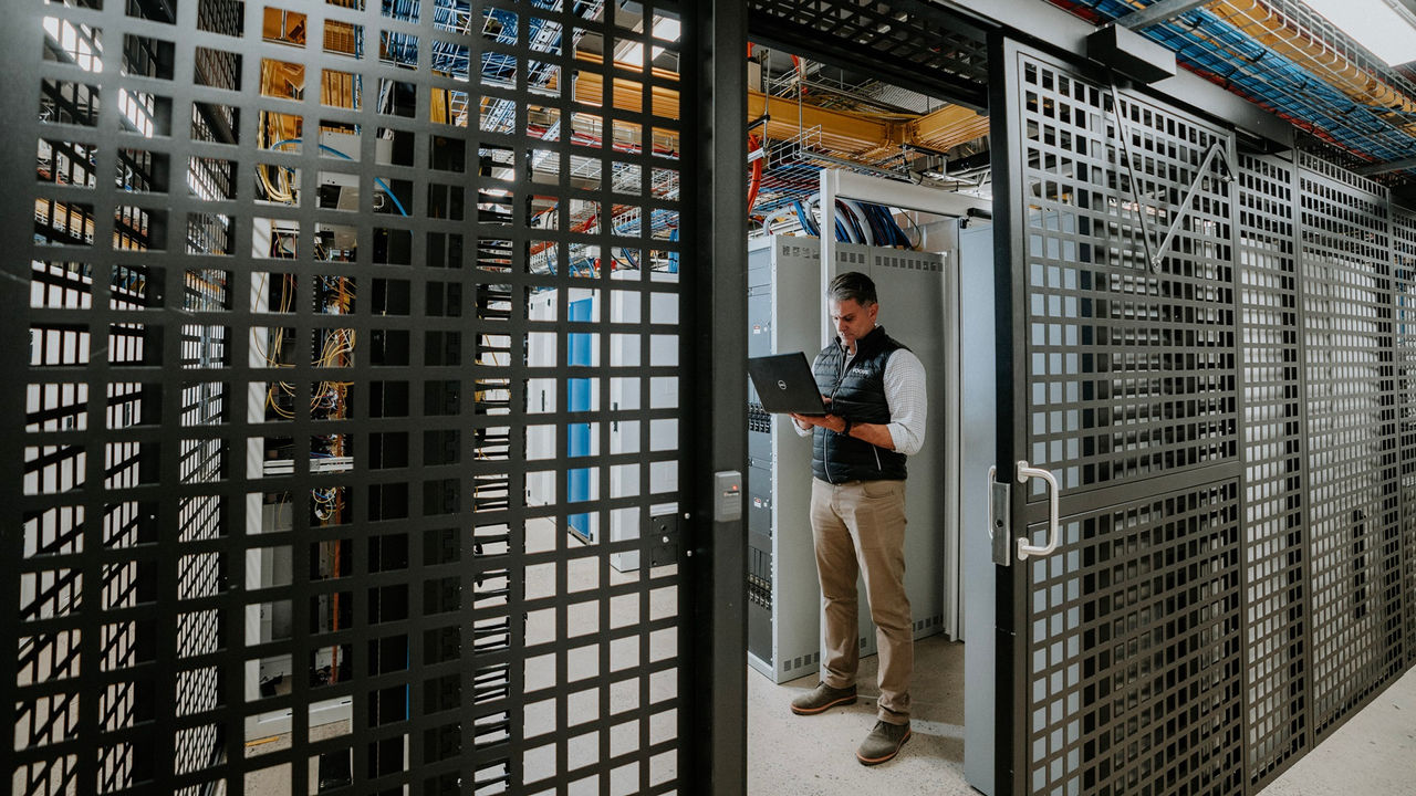 A technician stands inside a secure server room, working on a laptop with rows of network and server equipment visible, surrounded by metal security cages and overhead cable trays.