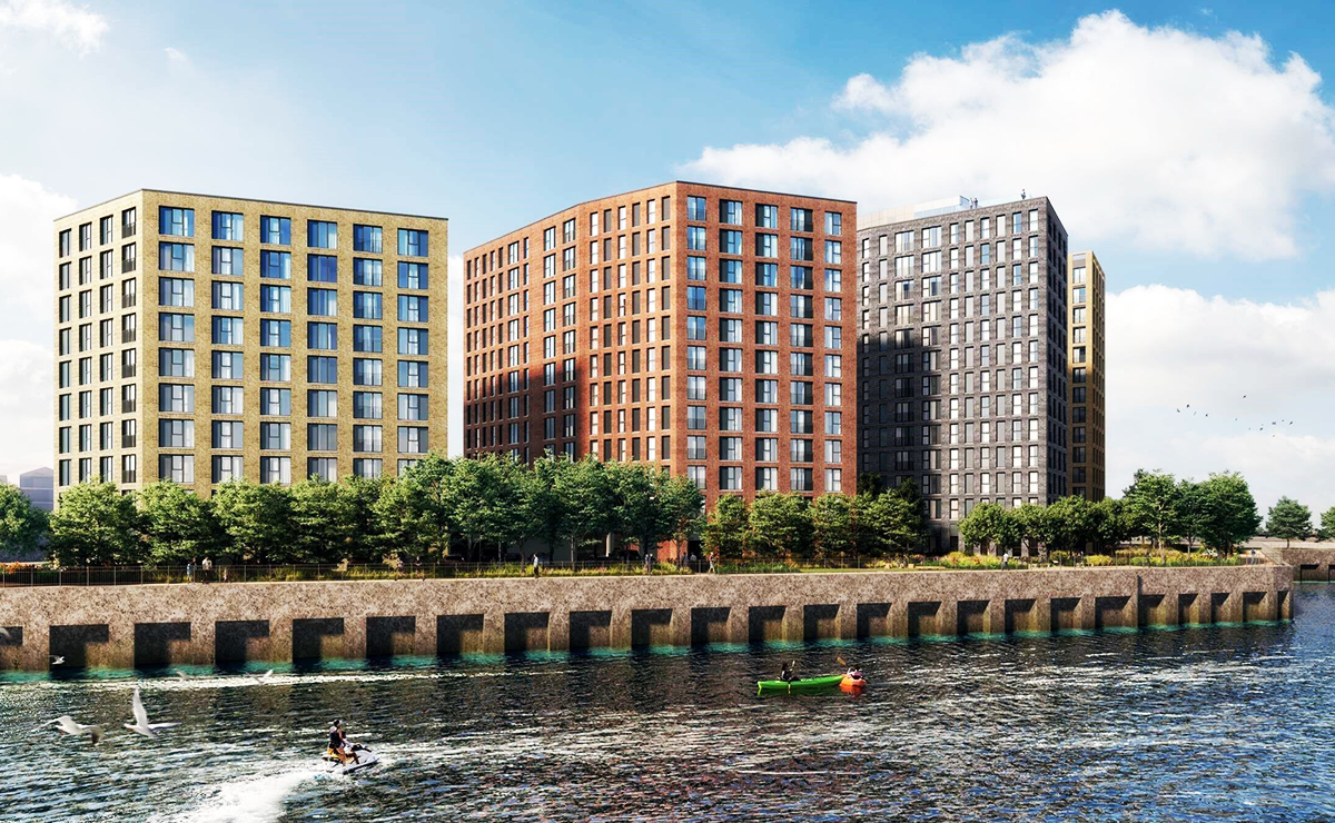Three contemporary residential apartment buildings in yellow, red, and dark gray brick facing a waterfront. Trees line the promenade, and people are kayaking and jet skiing in the water under a bright blue sky with scattered clouds.