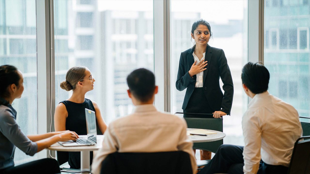 A young Indian Asian woman stands up in front of her diverse team and is leading a meeting, training or presentation in their office during the daytime. They are an ethnically diverse team.