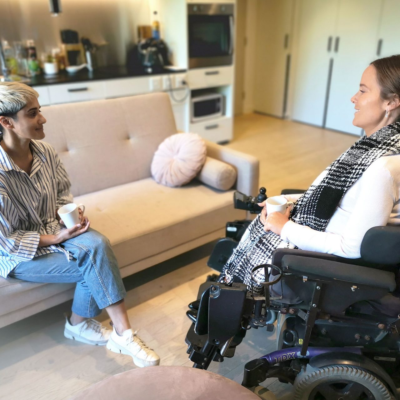 Two ladies having a coffee, one seated on a sofa, one in a wheelchair, in Australia's Specialist Disability Accommodation