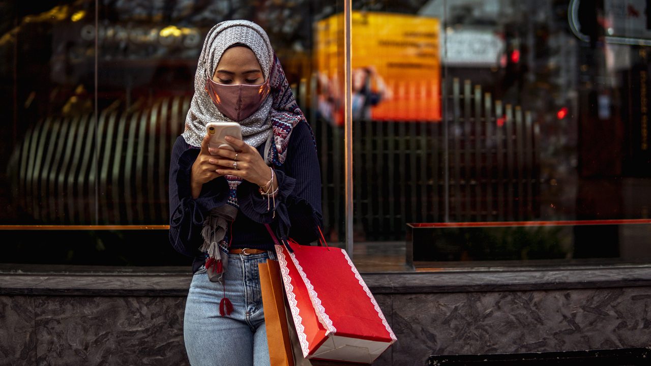 Portrait shot of a woman wearing a facemask whilst texting on a mobile phone and holding shopping bags.
