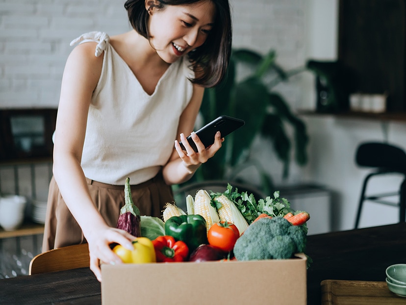 Beautiful smiling young Asian woman doing home delivery grocery shopping online with mobile app device on smartphone at home, with a box of colourful and fresh organic vegetables and fruits on the table