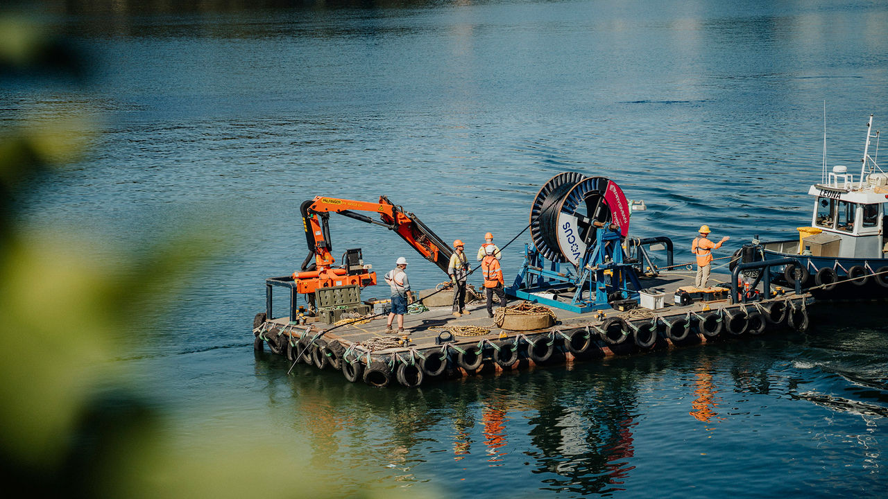 A group of workers in safety gear stand on a floating platform on a body of water, operating heavy machinery and equipment, including a large spool labeled "VOCUS." A small boat named "LEONA" is docked alongside the platform.