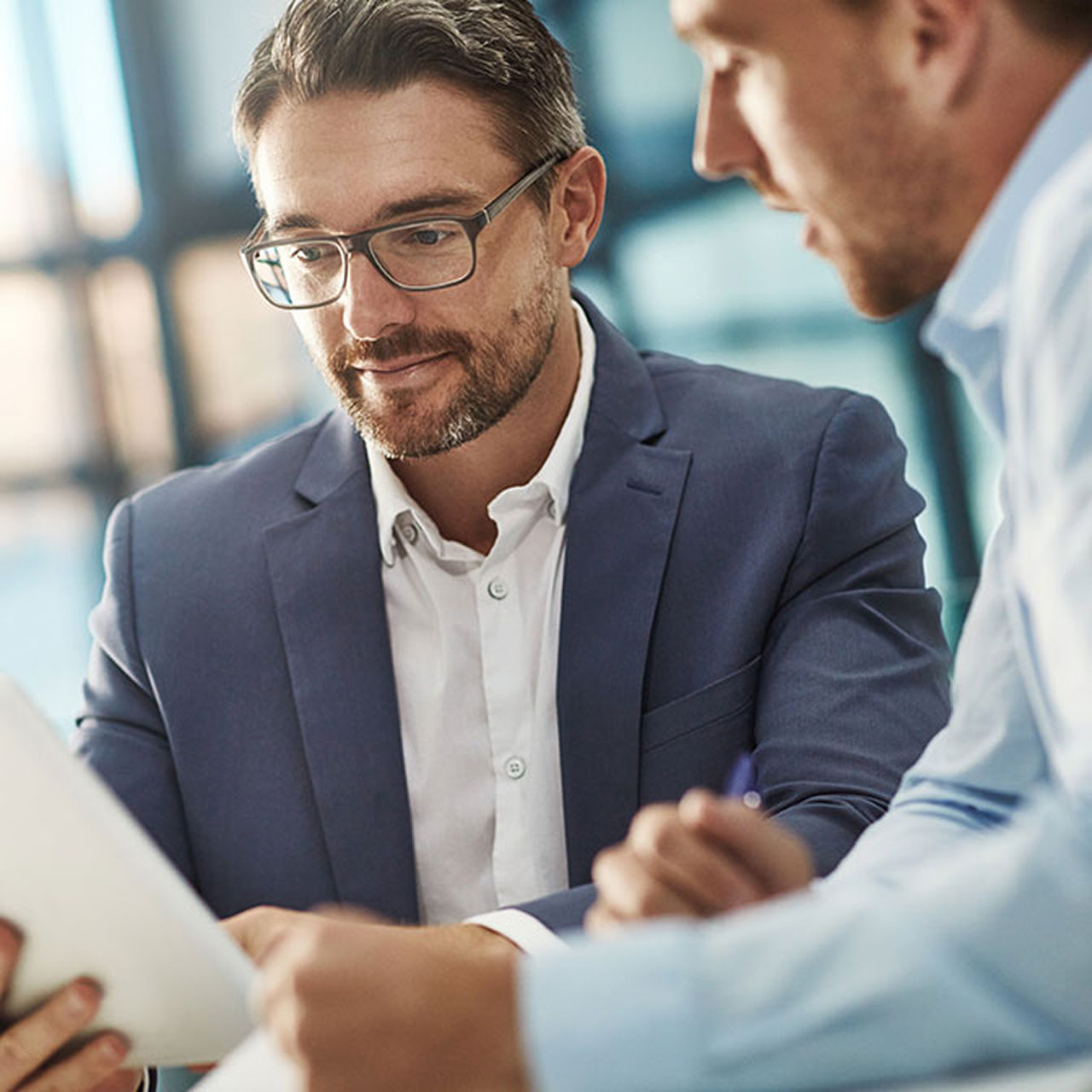 A business meeting with one person holding a tablet and another gesturing, set in a modern office with large windows.
