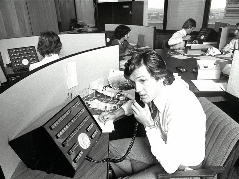Vintage office scene with employees working at desks with rotary phones and paperwork in a partitioned workspace.