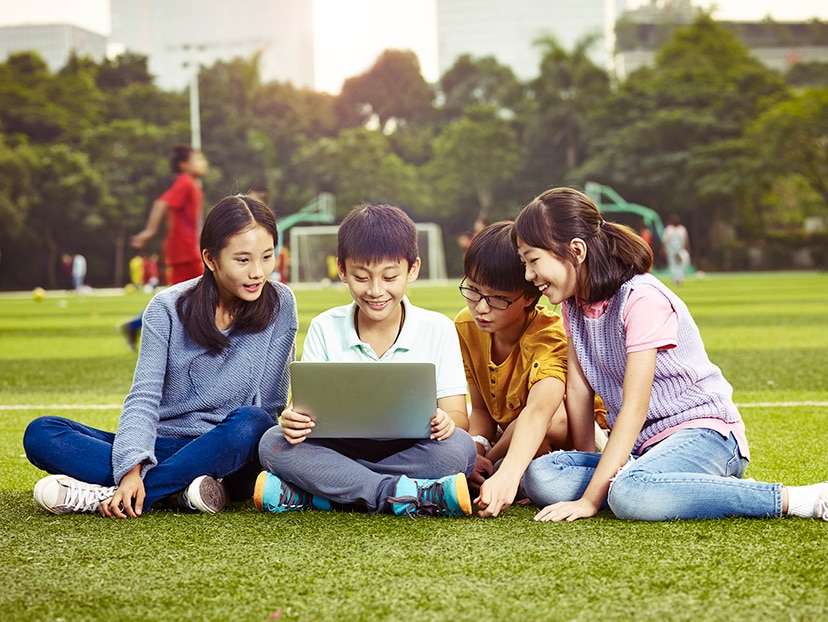 a group of asian elementary school children sitting on grass using laptop computer together.