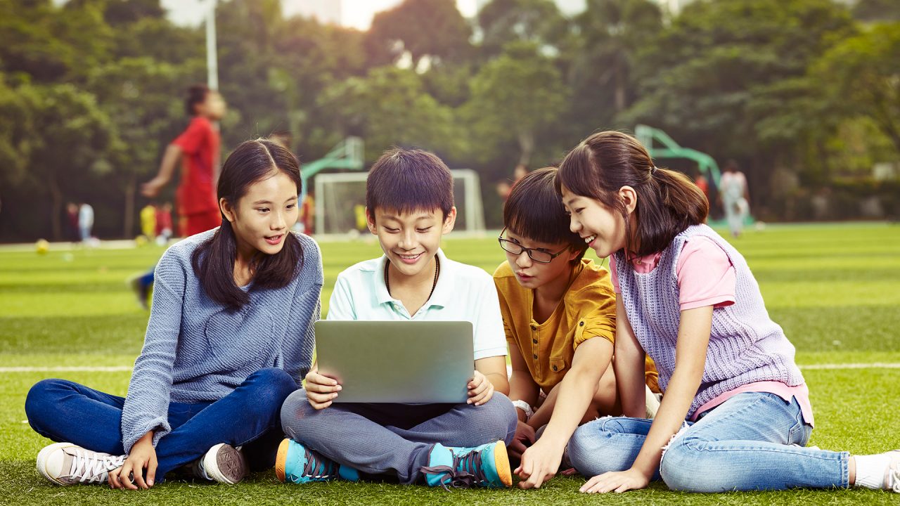 a group of asian elementary school children sitting on grass using laptop computer together.