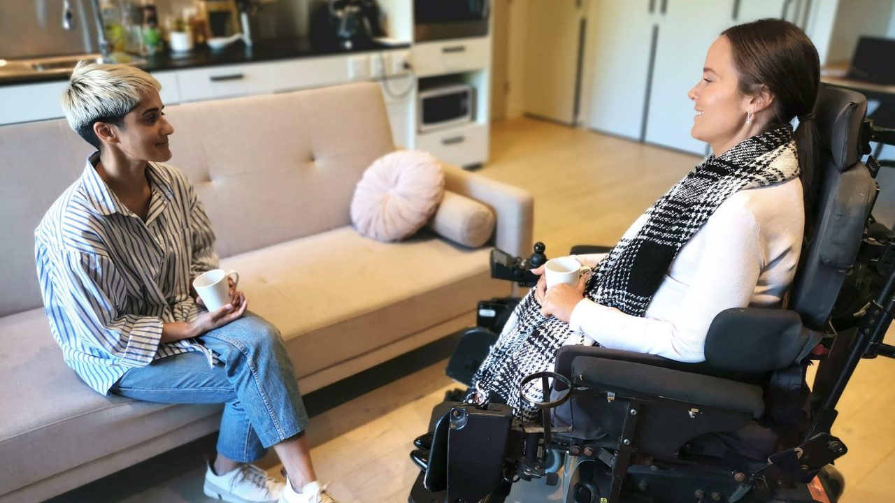 A woman sat on a pastel pink sofa speaking to another woman in a black wheelchair