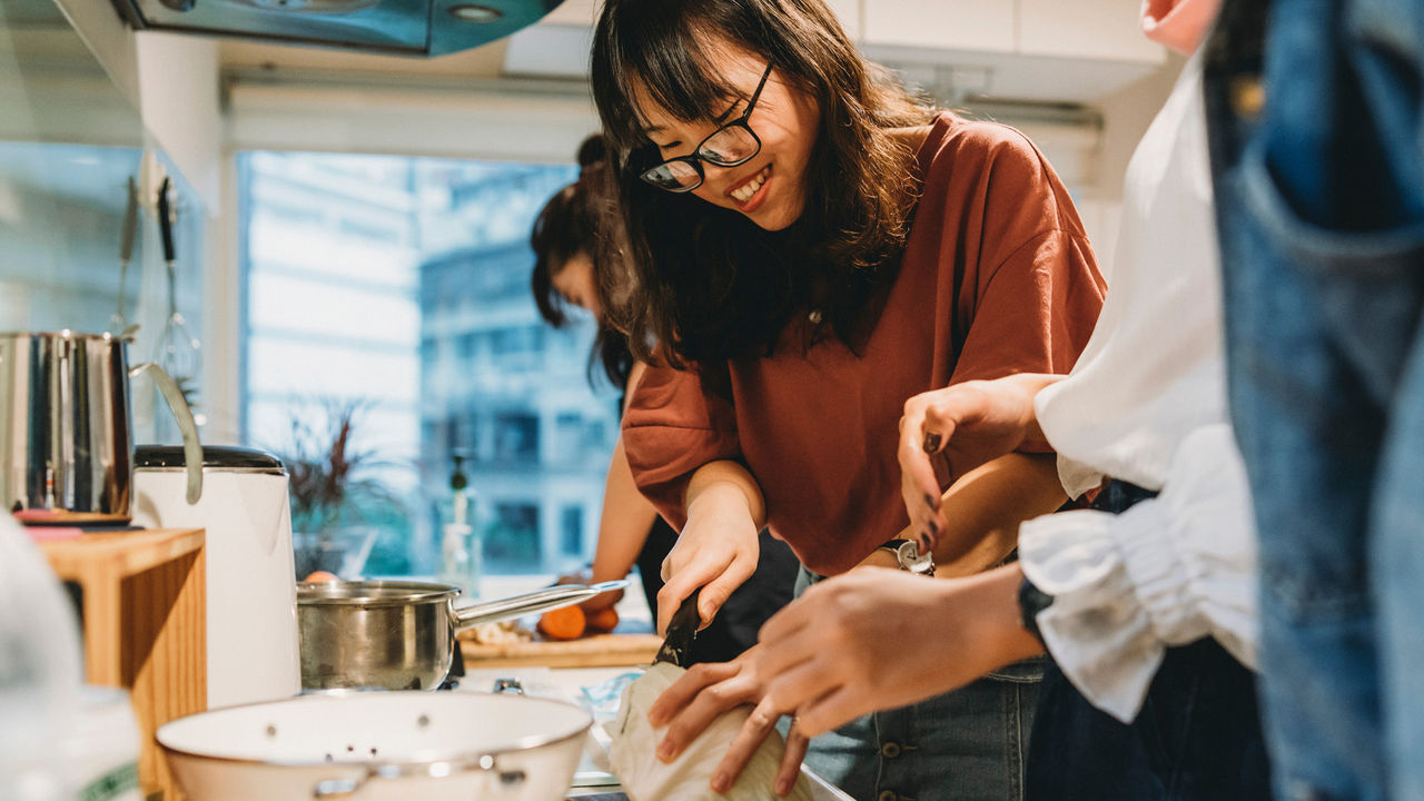 A group of people preparing food together in a kitchen, with one person chopping ingredients at the counter while others assist nearby.
