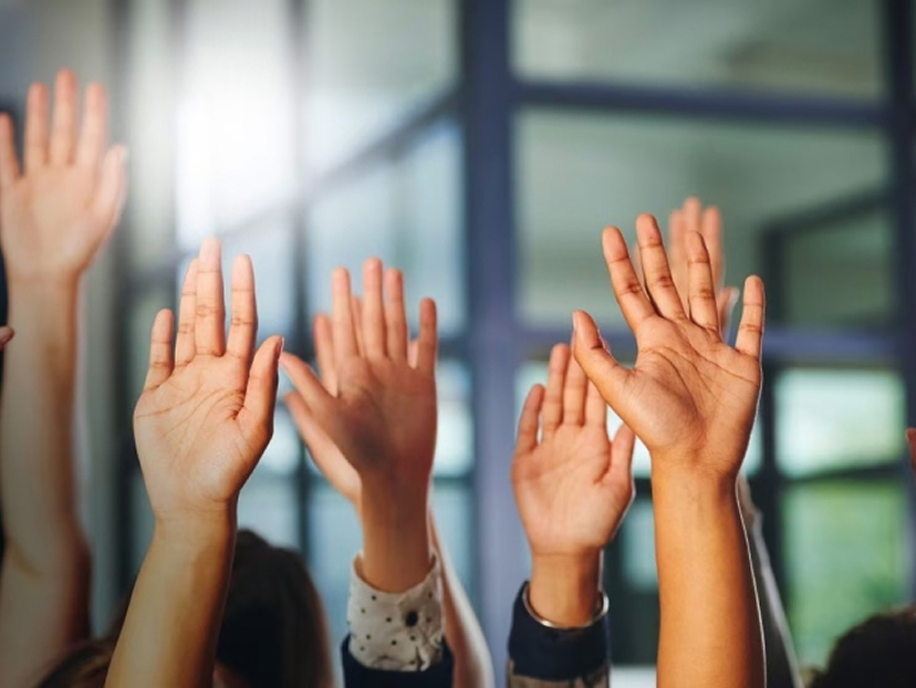 Multiple hands raised in the air, symbolizing participation or voting in a bright indoor setting.