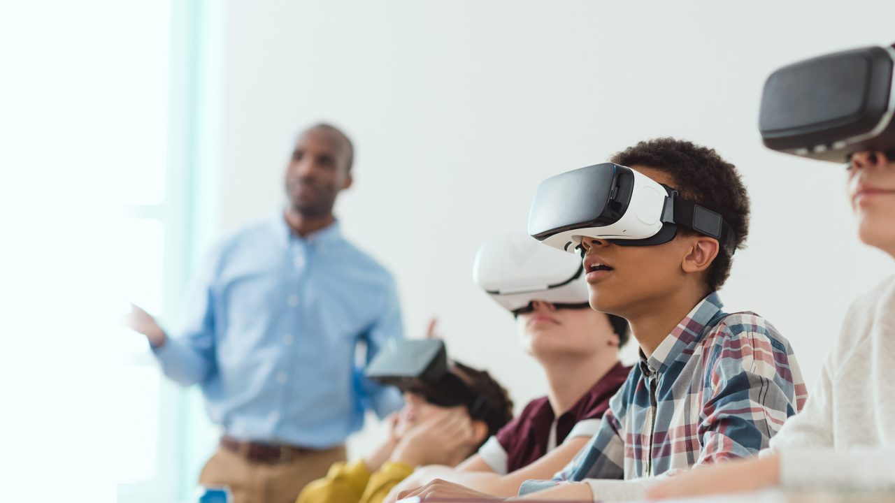 Multicultural school children using virtual reality headsets and teacher standing behind
