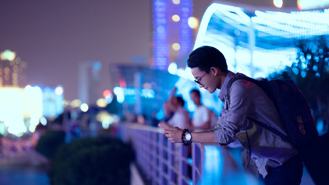 A young man leaning over a rail whilst simultaneously scrolling on his smart phone device