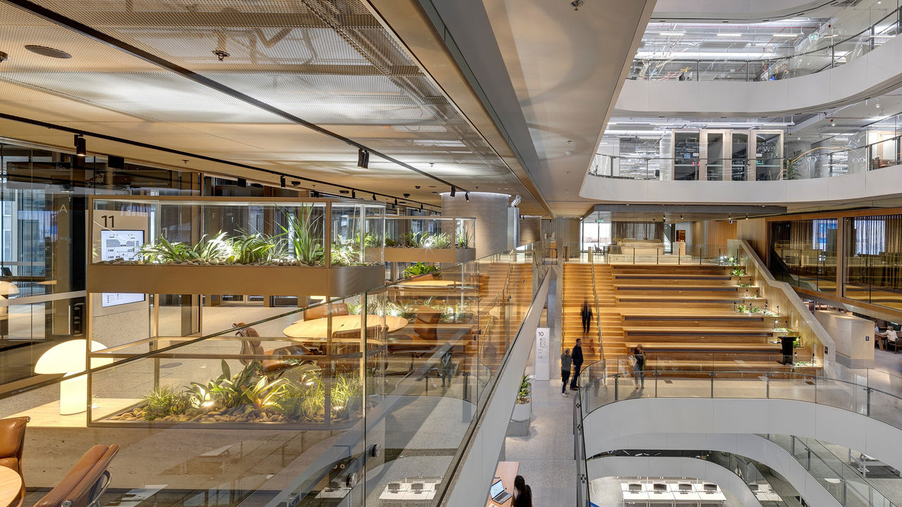 A modern office interior with open workspaces, glass partitions, large wooden steps for seating, indoor plants, and people working and walking.